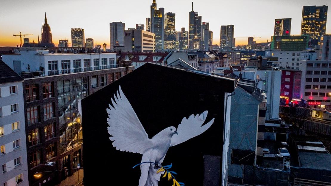 Auf eine Hausfassade wurde eine weiße Taube vor schwarzem Hintergrund gemalt. Sie trägt einen Ölzweig in ukrainischen Farben. Hinter dem Haus sieht man die Frankfurter Skyline.
