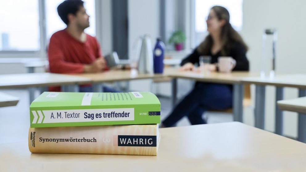 Ein kleiner Stapel an Deutschlern-Büchern, im Hintergrund zwei Menschen